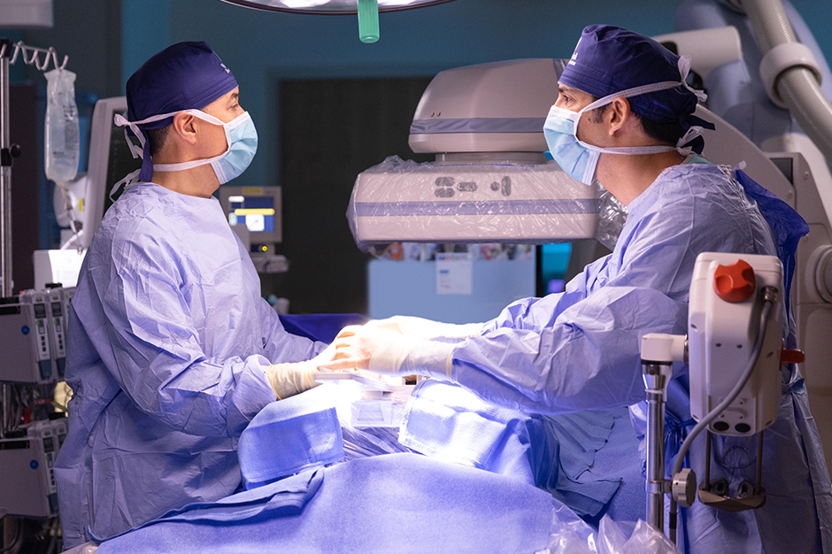 Two surgeons in blue scrubs, masks, and surgical caps perform a cardiac catheterization procedure together in an operating room
