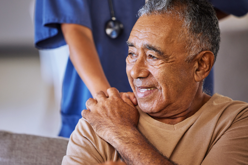 An older man smiles while holding his shoulder as a healthcare worker stands behind him